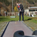 Eine Person, die im Freien Minigolf spielt, bereitet sich darauf vor, einen gelben Golfball auf einem schmalen Platz mit Bäumen und Hügeln im Hintergrund zu schlagen.