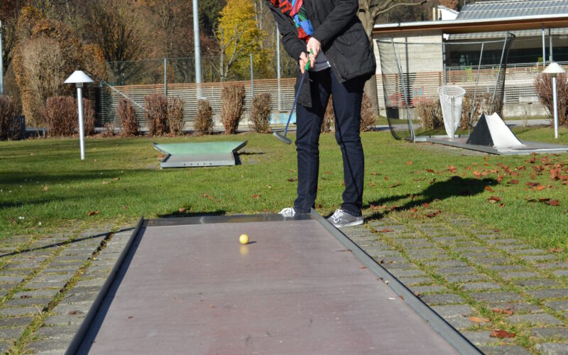 Eine Person, die im Freien Minigolf spielt, bereitet sich darauf vor, einen gelben Golfball auf einem schmalen Platz mit Bäumen und Hügeln im Hintergrund zu schlagen.