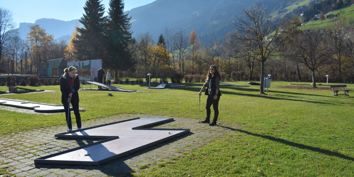 Zwei Frauen spielen Minigolf an einem sonnigen Tag in einem grasbewachsenen Park mit Bergen und Bäumen im Hintergrund.