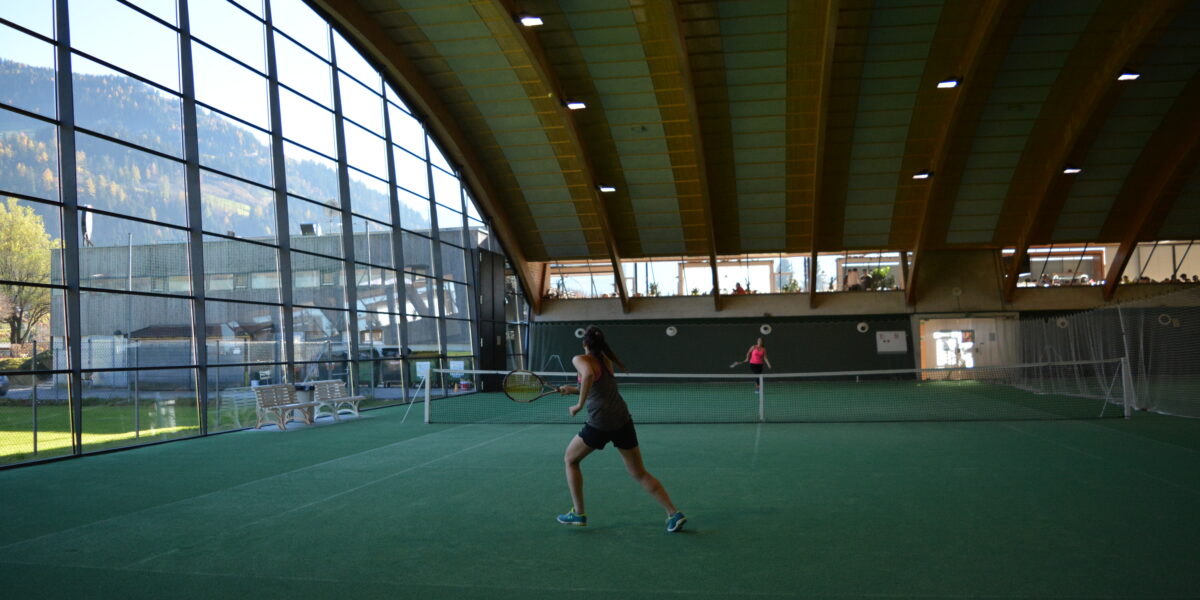 Zwei Personen spielen Tennis auf einem überdachten Platz mit einem geschwungenen Holzdach und großen Fenstern, die den Blick auf die Berge freigeben.