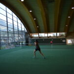 Zwei Personen spielen Tennis auf einem überdachten Platz mit einem geschwungenen Holzdach und großen Fenstern, die den Blick auf die Berge freigeben.