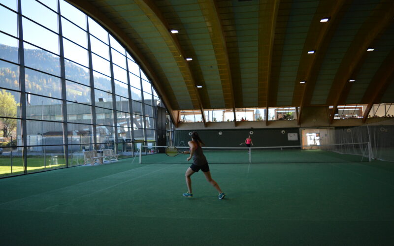 Zwei Personen spielen Tennis auf einem überdachten Platz mit einem geschwungenen Holzdach und großen Fenstern, die den Blick auf die Berge freigeben.