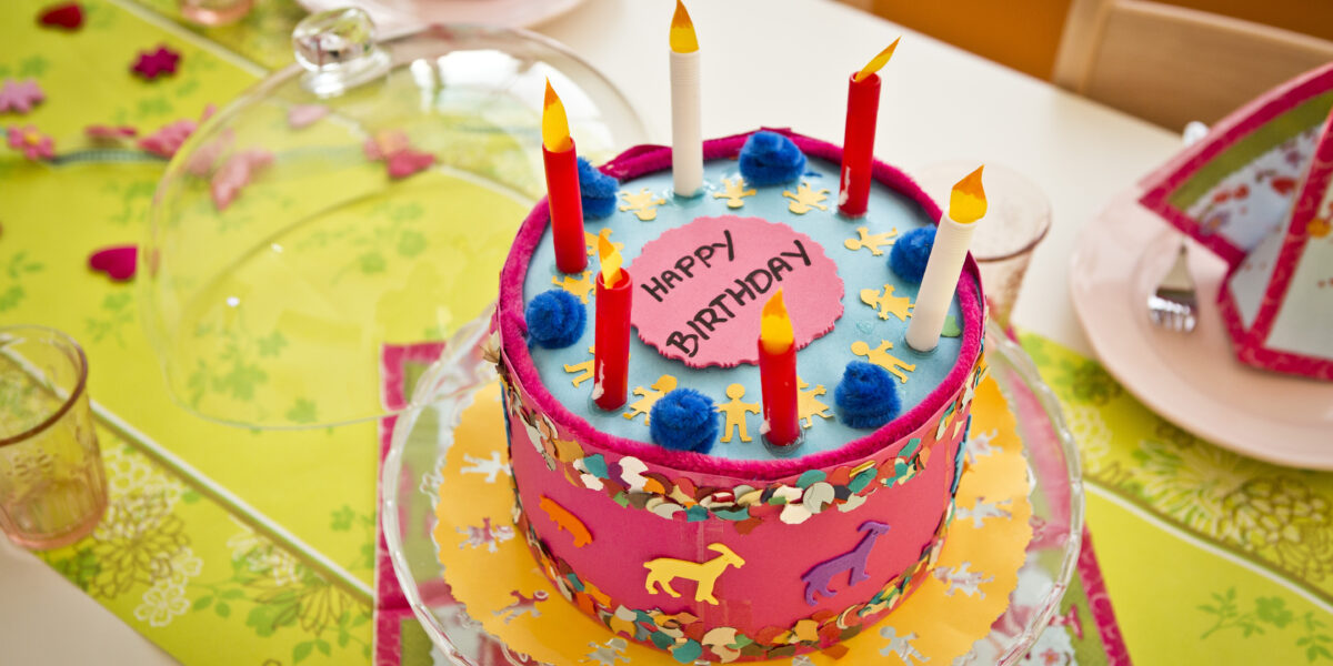Ein bunter Geburtstagskuchen mit sechs unbeleuchteten Kerzen und der Aufschrift "Happy Birthday" steht auf einem dekorierten Tisch mit Partyhüten, Gläsern und Servietten.