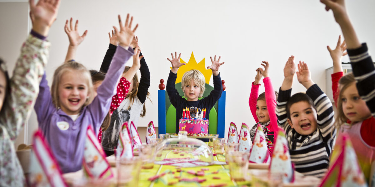 Kinder sitzen um einen Tisch mit Partyhüten und Dekorationen und heben feierlich die Hände, während ein Kind in der Mitte vor einer Geburtstagstorte sitzt.