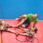Ein Erwachsener beaufsichtigt zwei Kinder an einer Kletterwand und hilft ihnen mit Seilen, während sie auf einer rot gepolsterten Matte sitzen.