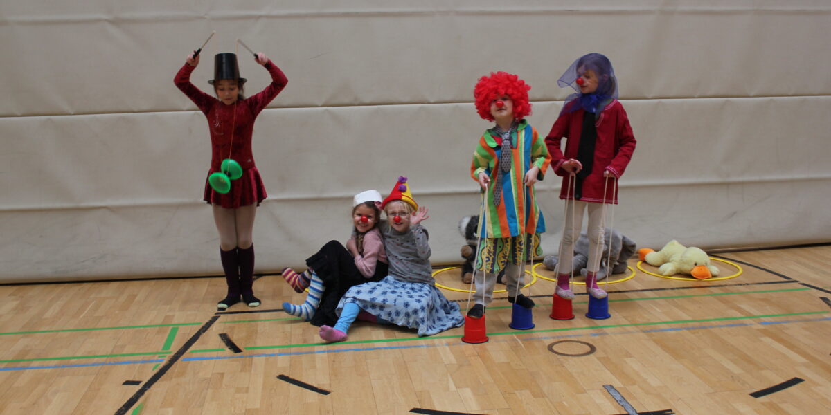 Fünf Kinder in bunten Zirkuskostümen treten in einer Turnhalle mit Requisiten wie Clownsnasen, Hüten, Stelzen und Jonglierstäben auf.