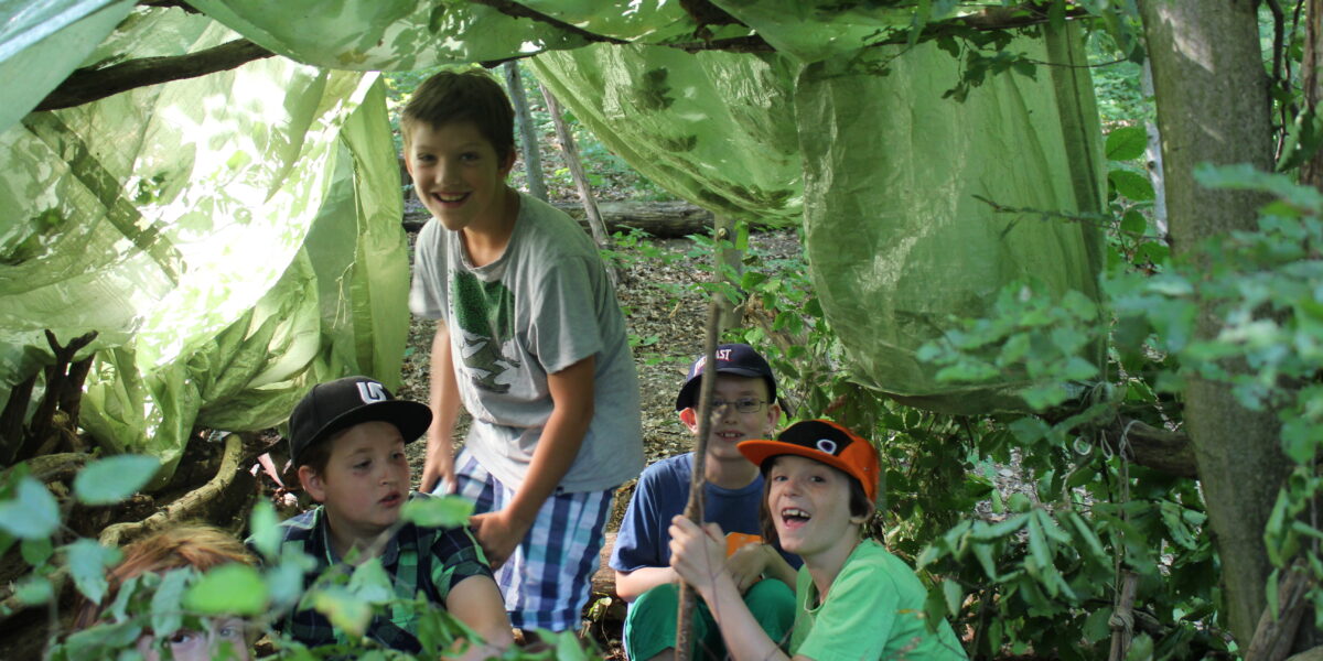 Vier Kinder sitzen und eines steht in einem aus Stöcken und grünen Planen gebauten Fort in einem Waldgebiet, umgeben von Bäumen und Laub.