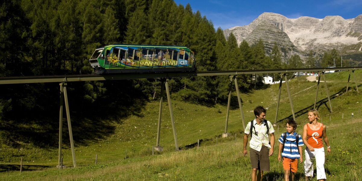 Eine vierköpfige Familie geht auf einem grasbewachsenen Hügel spazieren, während über ihnen eine Einschienenbahn vorbeifährt, mit Wald und Bergen im Hintergrund.