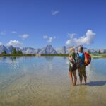 Zwei Wanderer stehen in flachem, klarem Wasser in der Nähe eines Sees, umgeben von Bergen und einem blauen Himmel mit vereinzelten Wolken.