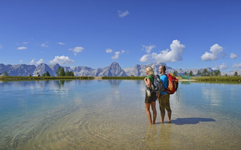 Zwei Wanderer stehen in flachem, klarem Wasser in der Nähe eines Sees, umgeben von Bergen und einem blauen Himmel mit vereinzelten Wolken.
