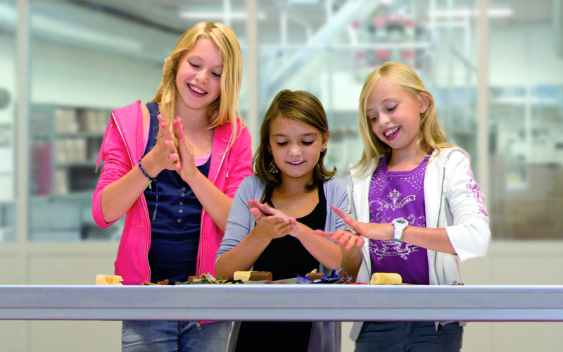 Drei Mädchen stehen lächelnd an einem Tisch und formen in einem hell erleuchteten Raum mit ihren Händen Ton oder Teig.