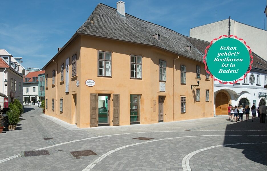 Ein historisches, zweistöckiges, beigefarbenes Gebäude mit einem Schild mit der Aufschrift "Beethovenhaus" steht an einer Straßenecke in Baden, Österreich, unter klarem Himmel. Eine Gruppe von Menschen geht in der Nähe spazieren.