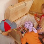 Vier Kinder schauen in eine offene Holztruhe, die mit verschiedenen kleinen Spielsachen und Gegenständen gefüllt ist.