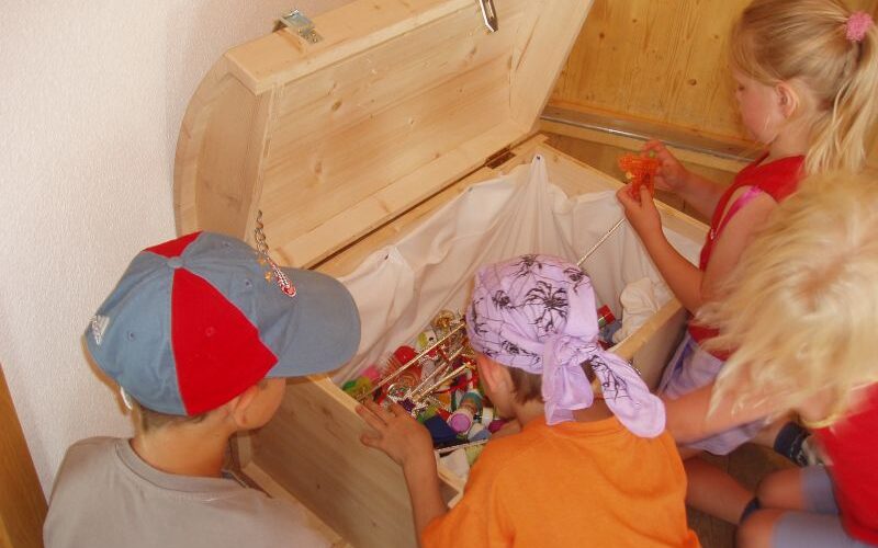 Vier Kinder schauen in eine offene Holztruhe, die mit verschiedenen kleinen Spielsachen und Gegenständen gefüllt ist.