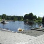 Auf einer von Bäumen umgebenen Holzterrasse am Seeufer kann man sich entspannen und schwimmen, während eine Person ins Wasser springt und es dabei ordentlich krachen lässt.