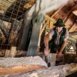 Ein Mann in traditioneller Kleidung sägt mit einer großen Vertikalsäge einen Stamm in einer rustikalen Holzwerkstatt.