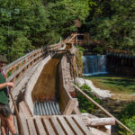 Ein Mann steht auf einem Holzsteg neben einem Bach mit einem kleinen Wasserfall, umgeben von dichtem grünen Wald.