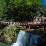 Eine Holzbrücke führt über einen kleinen Wasserfall in einem bewaldeten Gebiet unter blauem Himmel.