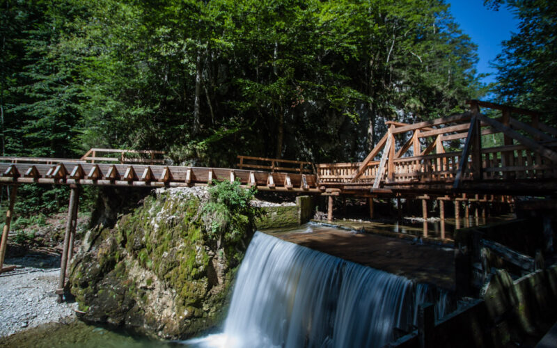Eine Holzbrücke führt über einen kleinen Wasserfall in einem bewaldeten Gebiet unter blauem Himmel.