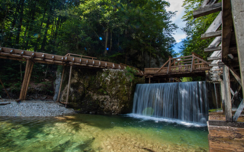 Unter einem hölzernen Steg, der von Wald umgeben ist, fließt ein kleiner Wasserfall, in dem sich klares Wasser sammelt und das Sonnenlicht durch die Bäume fällt.