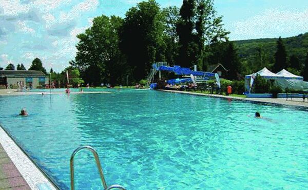 Freibad mit blauem Wasser, ein paar schwimmende Leute, Wasserrutsche im Hintergrund, Bäume, Hügel und Zelte unter einem teilweise bewölkten Himmel sichtbar.