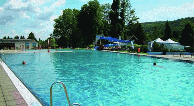 Freibad mit blauem Wasser, ein paar schwimmende Leute, Wasserrutsche im Hintergrund, Bäume, Hügel und Zelte unter einem teilweise bewölkten Himmel sichtbar.