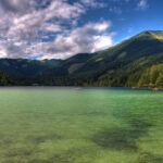 Klarer See mit grünlichem Wasser, umgeben von bewaldeten Bergen unter einem teilweise bewölkten Himmel. Kleine Boote und Gebäude sind entlang der Uferlinie zu sehen.