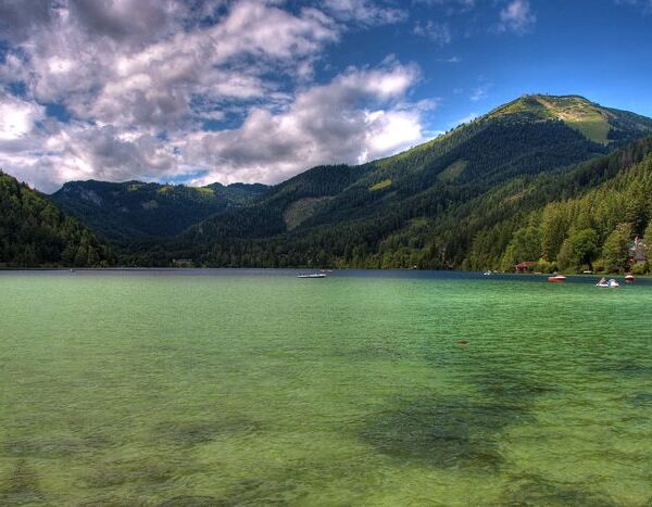 Klarer See mit grünlichem Wasser, umgeben von bewaldeten Bergen unter einem teilweise bewölkten Himmel. Kleine Boote und Gebäude sind entlang der Uferlinie zu sehen.