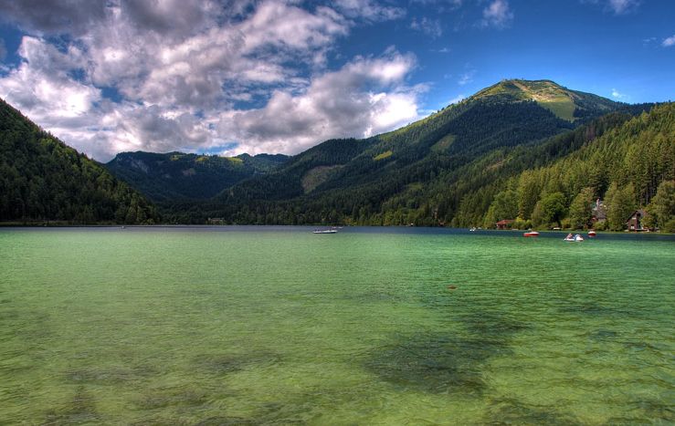 Klarer See mit grünlichem Wasser, umgeben von bewaldeten Bergen unter einem teilweise bewölkten Himmel. Kleine Boote und Gebäude sind entlang der Uferlinie zu sehen.