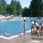 Kinder versammeln sich am Rande eines Freibads, während andere schwimmen; im Hintergrund sind Bäume und Menschen zu sehen.