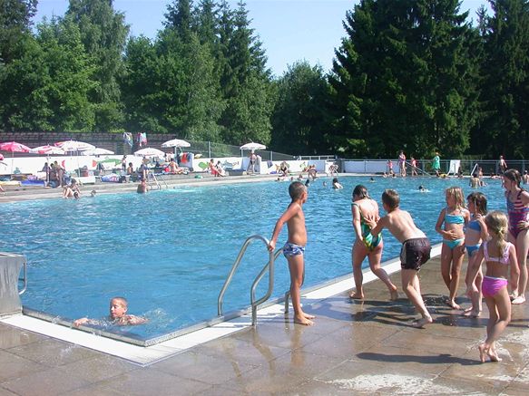 Kinder versammeln sich am Rande eines Freibads, während andere schwimmen; im Hintergrund sind Bäume und Menschen zu sehen.