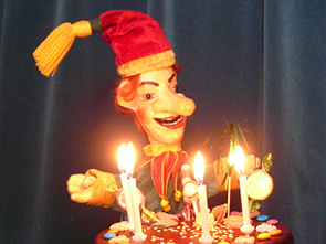 Eine Marionette mit rotem Hut und gelber Quaste steht hinter einer Torte mit brennenden Kerzen und bunten Dekorationen.