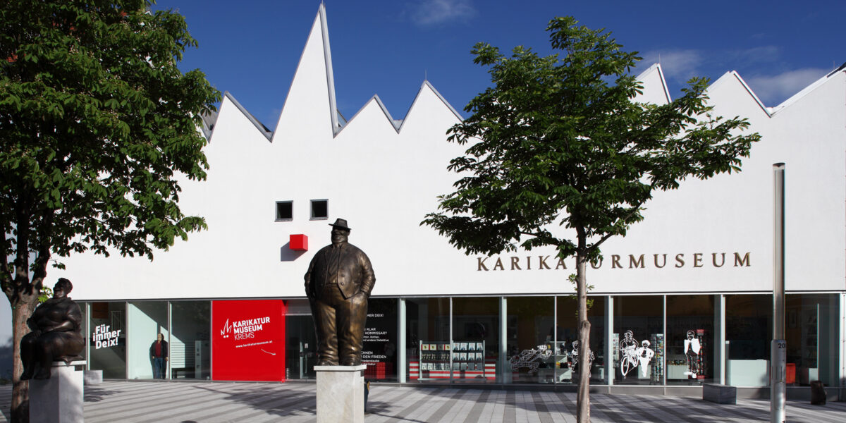 Weißes, modernes Gebäude mit gezacktem Dach und der Aufschrift "Karikaturmuseum"; Statuen auf Sockeln und Bäume säumen den gestreiften Platz davor unter einem klaren blauen Himmel.