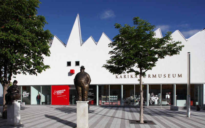 Weißes, modernes Gebäude mit gezacktem Dach und der Aufschrift "Karikaturmuseum"; Statuen auf Sockeln und Bäume säumen den gestreiften Platz davor unter einem klaren blauen Himmel.