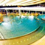 Hallenbadkomplex mit geschwungenen Becken, schwimmenden und spielenden Menschen, Liegestühlen entlang großer Fenster und Oberlicht.