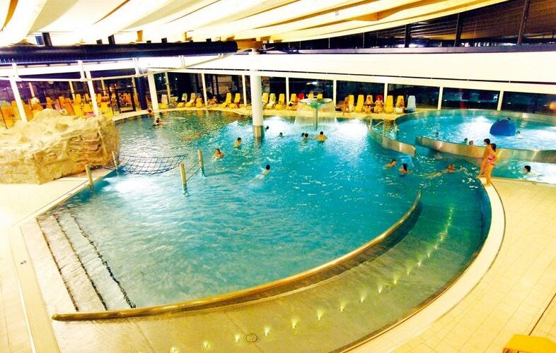 Hallenbadkomplex mit geschwungenen Becken, schwimmenden und spielenden Menschen, Liegestühlen entlang großer Fenster und Oberlicht.