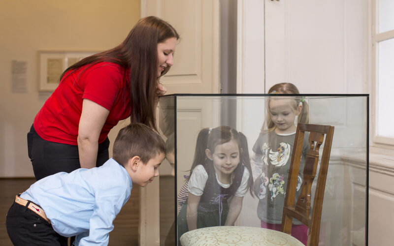 Eine Frau und drei kleine Kinder betrachten einen antiken Stuhl, der in einem Museum hinter Glas ausgestellt ist.
