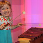 Ein kleines Kind lächelt, während es auf einem hölzernen Xylophon mit zwei Schlägeln spielt und vor einem rosa und lila Hintergrund steht.