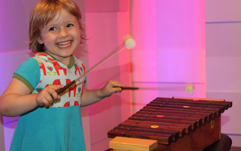 Ein kleines Kind lächelt, während es auf einem hölzernen Xylophon mit zwei Schlägeln spielt und vor einem rosa und lila Hintergrund steht.