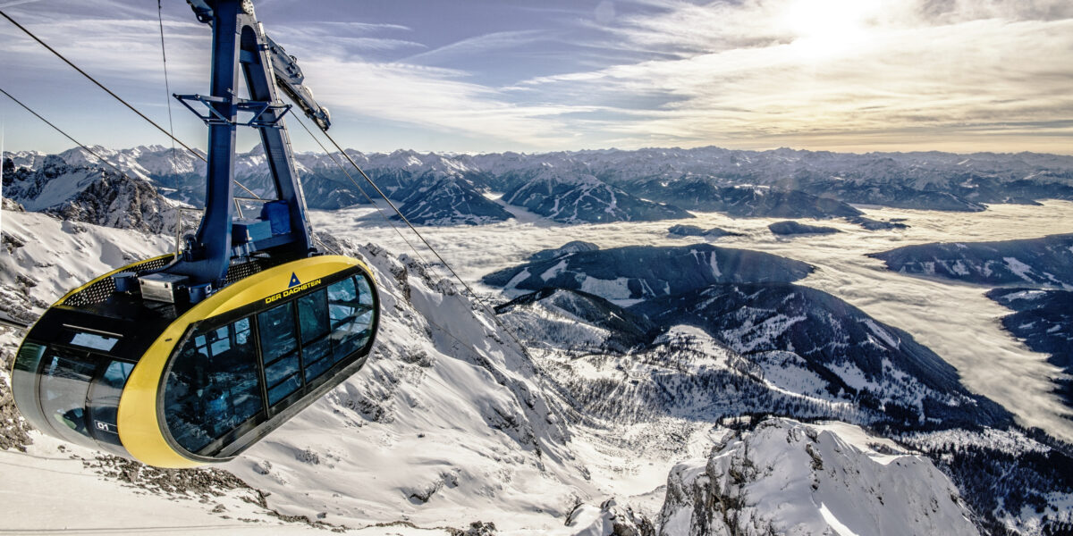 Eine gelb-schwarze Seilbahn erklimmt verschneite Berghänge unter einem klaren Himmel, mit fernen Berggipfeln und einem sonnenbeschienenen Tal darunter.