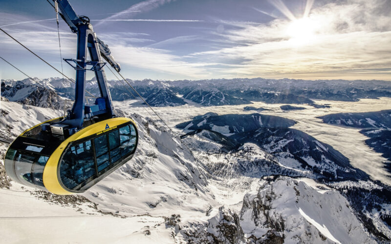 Eine gelb-schwarze Seilbahn erklimmt verschneite Berghänge unter einem klaren Himmel, mit fernen Berggipfeln und einem sonnenbeschienenen Tal darunter.