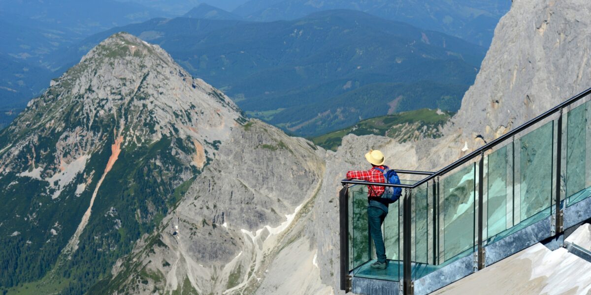 Eine Person mit Hut steht auf einer gläsernen Aussichtsplattform, von der aus man an einem klaren Tag Berge und Täler überblicken kann.