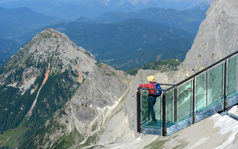 Eine Person mit Hut steht auf einer gläsernen Aussichtsplattform, von der aus man an einem klaren Tag Berge und Täler überblicken kann.