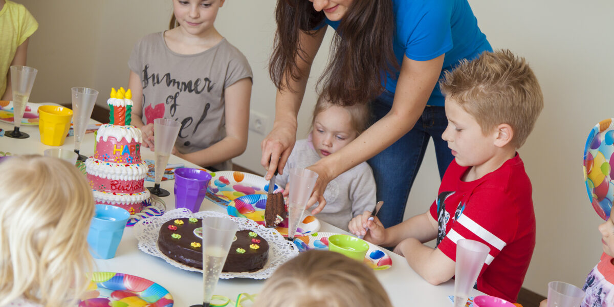 Eine Frau schneidet einen Schokoladenkuchen an einem Tisch an, an dem mehrere Kinder sitzen, umgeben von Geburtstagsdekorationen und bunten Tellern.