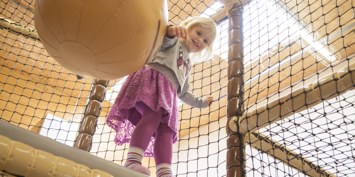Ein Kleinkind steht auf einem gepolsterten Podest in einem Indoor-Spielbereich und hält sich an einem großen weichen Ball fest, der von Sicherheitsnetzen umgeben ist.