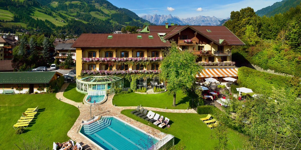 Ein Hotel mit Balkonen und Blumenkästen bietet einen Blick auf den Außenpool, den Whirlpool, die Sonnenliegen und die angelegten Gärten vor der Kulisse bewaldeter Hügel und entfernter Berge.