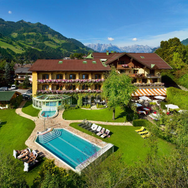 Ein Hotel mit Balkonen und Blumenkästen bietet einen Blick auf den Außenpool, den Whirlpool, die Sonnenliegen und die angelegten Gärten vor der Kulisse bewaldeter Hügel und entfernter Berge.