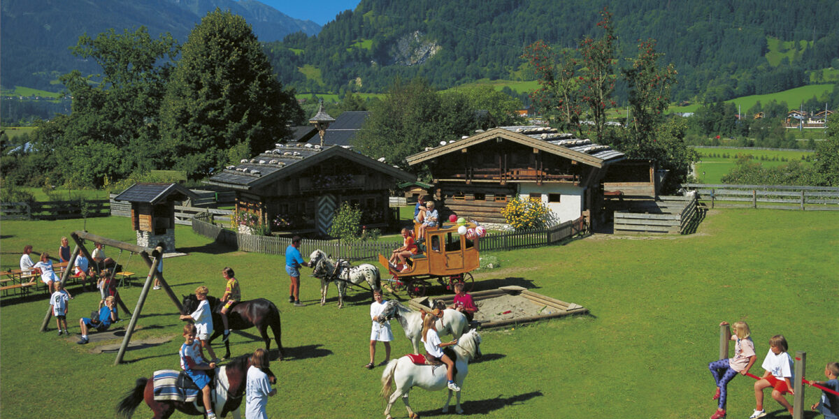 Kinder reiten auf Ponys, spielen auf Schaukeln und benutzen einen Sandkasten in einem grasbewachsenen Hof mit Holzhäusern und Bergen im Hintergrund an einem sonnigen Tag.