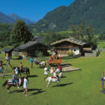 Kinder reiten auf Ponys, spielen auf Schaukeln und benutzen einen Sandkasten in einem grasbewachsenen Hof mit Holzhäusern und Bergen im Hintergrund an einem sonnigen Tag.