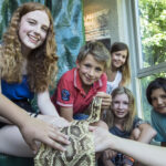 Fünf Kinder sitzen in einem Haus, lächeln und berühren eine große Schlange, die von einem Erwachsenen gehalten wird. Bäume und Sonnenlicht sind durch das Fenster hinter ihnen zu sehen.